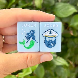May include: Two light blue square tiles with the word "JOKER" printed on them. One tile features a mermaid design, the other a sailor with a beard. The tiles are held against a backdrop of green foliage.