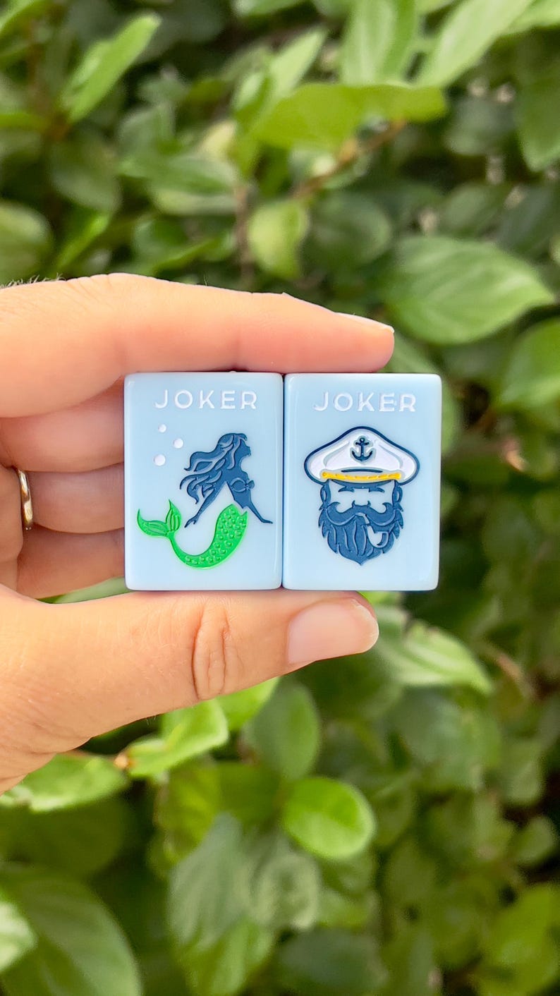 May include: Two light blue square tiles with the word "JOKER" printed on them. One tile features a mermaid design, the other a sailor with a beard. The tiles are held against a backdrop of green foliage.