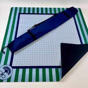 May include: A blue, green, and white striped beach mat with a matching carrying case. The mat features a light blue woven pattern in the center and a dark blue border. A logo is in the corner.
