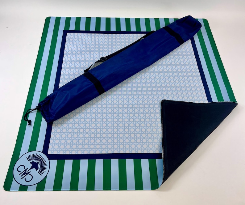 May include: A blue, green, and white striped beach mat with a matching carrying case. The mat features a light blue woven pattern in the center and a dark blue border. A logo is in the corner.