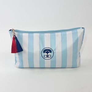 May include: A rectangular cosmetic bag with vertical light blue and white stripes. It features a gold zipper, a red and navy blue tassel, and a circular logo with the initials "CMO".