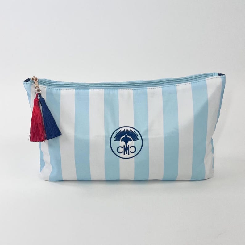 May include: A rectangular cosmetic bag with vertical light blue and white stripes. It features a gold zipper, a red and navy blue tassel, and a circular logo with the initials "CMO".