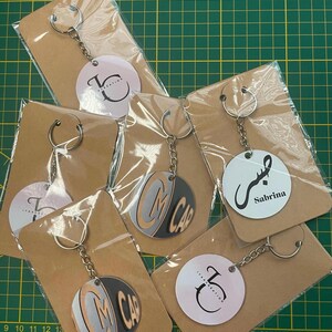Peut inclure: Cinq porte-clés avec des étiquettes circulaires rondes, blanches et noires. Les étiquettes présentent différents motifs, notamment un logo, un nom et de la calligraphie arabe. Les porte-clés sont emballés dans des sachets en plastique transparent et du papier brun.