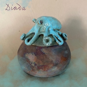 Op de afbeelding: Een keramieken pot met een deksel voorzien van een gebeeldhouwde lichtblauwe octopus met donkere stippen. De pot heeft een gevlekte, veelkleurige afwerking in tinten bruin, koper en donkerblauw. Het woord Diada staat in de linkerbovenhoek.