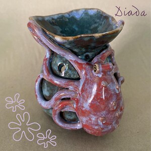 May include: A ceramic oil burner featuring a sculpted red octopus with textured tentacles wrapping around a dark teal, glossy vessel. The piece includes circular cutouts and the word Diada written in the upper right corner with three flower icons.