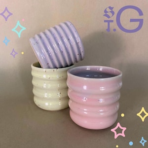 Könnte beinhalten: Drei Keramikbecher mit gerippter, wellenförmiger Textur. Ein Becher ist hellviolett mit dunklen Sprenkeln, einer ist blassgelb mit dunklen Sprenkeln und einer ist einfarbig hellrosa. Der Text ST.G erscheint oben rechts mit Sternengrafiken.