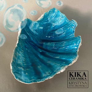 Könnte beinhalten: Eine muschelförmige Keramikschale mit einer glänzenden, leuchtend blauen Glasur und strukturierten Rillen. Das Innere zeigt gemalte weiße Blasenmuster. Ein schwarzes quadratisches Logo enthält den Text KIKA CERAMIKA KRYSTYNA STECZKOWSKA.