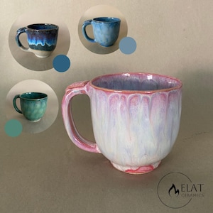 Pode incluir: Uma caneca de cerâmica com acabamento em esmalte escorrido rosa, branco e azul claro e uma alça resistente. Três inserções circulares mostram canecas adicionais em variações de azul, azul escuro e verde. O logotipo ELAT CERAMICS está no canto.
