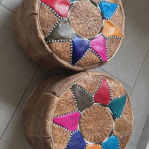 Puede incluir: Dos pufs redondos de cuero marrón con patrones geométricos coloridos en la parte superior. Cada puf tiene un diseño floral central en relieve rodeado de parches triangulares en varios colores, incluyendo azul, rosa y naranja. Los lados tienen un patrón floral en relieve.
