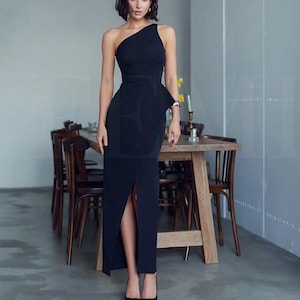 Puede incluir: Una mujer con un vestido negro de maxi largo con un hombro descubierto y una abertura en el muslo. El vestido tiene un corpiño ajustado y una falda fluida. La mujer está de pie frente a una mesa de madera con sillas.