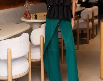 May include: A black strapless top with layered ruffles paired with emerald green wide-leg pants. The pants have a wrap-style design. The outfit is worn with black pointed-toe heels. The background shows a restaurant setting.