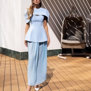 May include: A light blue two-piece outfit featuring a top with short sleeves and a peplum waist, paired with a matching floor-length skirt. The outfit is accessorized with white sunglasses and white pointed-toe heels. The background includes a wooden deck and a hanging chair.