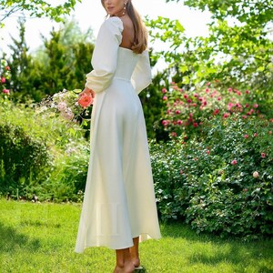 Pu&ograve; includere: Un abito maxi bianco con scollo quadrato e maniche lunghe. L'abito ha un corpetto aderente e una gonna fluida. La modella &egrave; in piedi in un giardino con erba verde e fiori rosa.