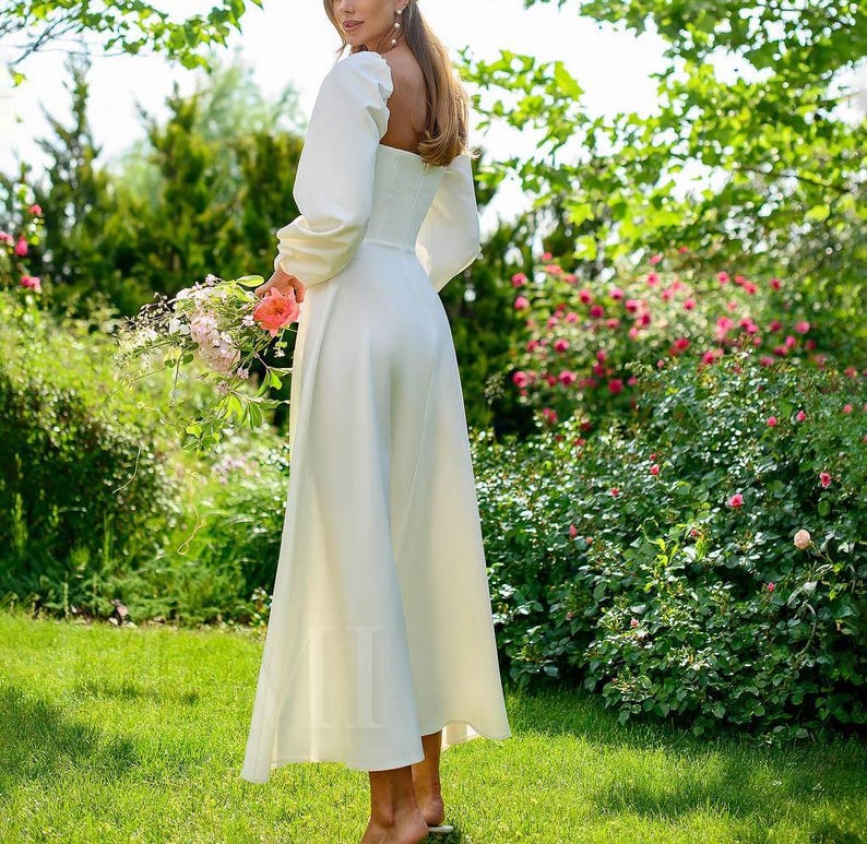 Pu&ograve; includere: Un abito maxi bianco con scollo quadrato e maniche lunghe. L'abito ha un corpetto aderente e una gonna fluida. La modella &egrave; in piedi in un giardino con erba verde e fiori rosa.