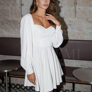 Pu&ograve; includere: Un abito bianco scollato con maniche lunghe e un corpetto aderente. L'abito ha una gonna svasata e una scollatura a cuore.