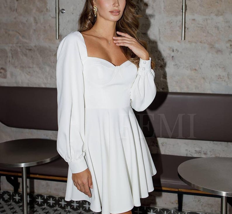 Pu&ograve; includere: Un abito bianco scollato con maniche lunghe e un corpetto aderente. L'abito ha una gonna svasata e una scollatura a cuore.