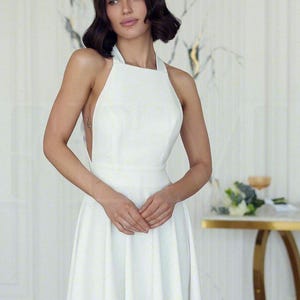 May include: A white halter-neck dress with a fitted bodice and a flowing skirt. The dress has a simple, elegant design, perfect for a special occasion. The model is wearing the dress in a well-lit room.