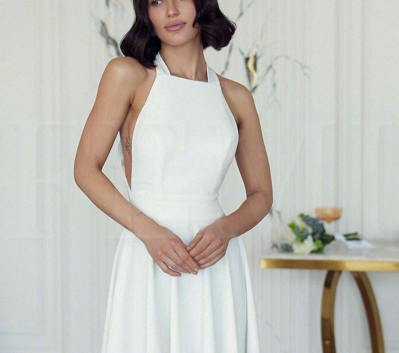 May include: A white halter-neck dress with a fitted bodice and a flowing skirt. The dress has a simple, elegant design, perfect for a special occasion. The model is wearing the dress in a well-lit room.