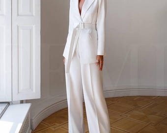 May include: A white blazer with a matching pair of wide-leg pants. The blazer has a notched lapel, a single button closure, and a belt at the waist. The pants have a straight leg and a wide, flowing silhouette.