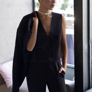 May include: A woman's black jumpsuit with a V-neck top and tailored pants. A black blazer is draped over the shoulders. A pearl necklace and earrings complete the look. The outfit is suitable for formal occasions.
