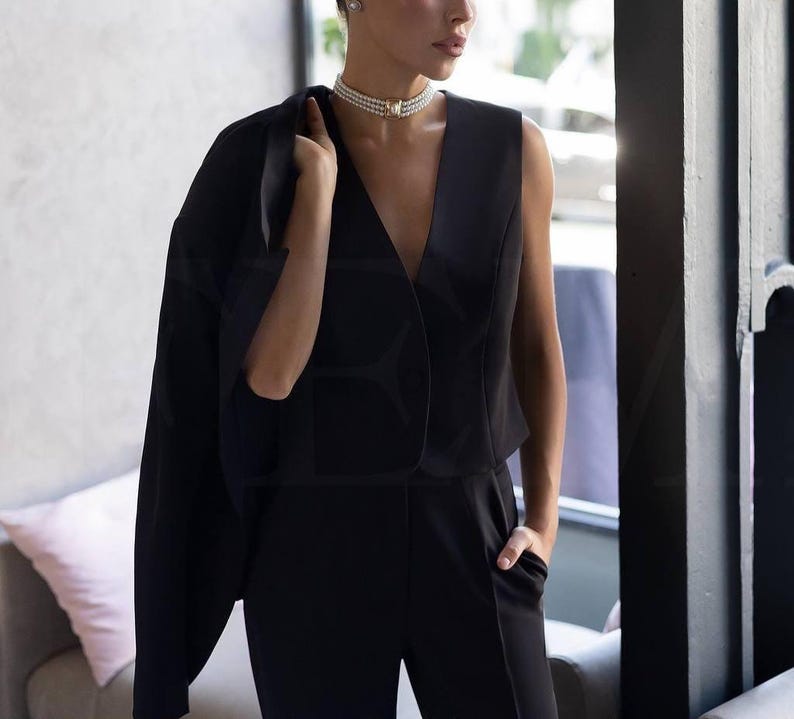 May include: A woman's black jumpsuit with a V-neck top and tailored pants. A black blazer is draped over the shoulders. A pearl necklace and earrings complete the look. The outfit is suitable for formal occasions.