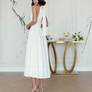 May include: A white halter-neck midi dress with a flowing skirt and a long, elegant tie at the back. The dress is paired with high-heeled sandals. A gold-framed table with a marble top and decorative elements is in the background.