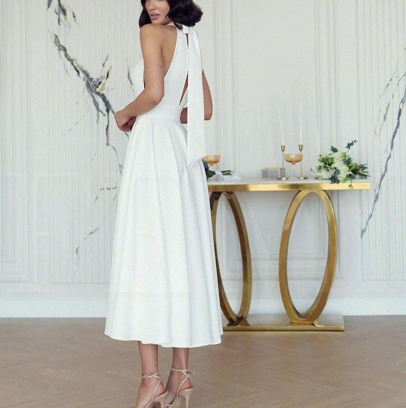 May include: A white halter-neck midi dress with a flowing skirt and a long, elegant tie at the back. The dress is paired with high-heeled sandals. A gold-framed table with a marble top and decorative elements is in the background.