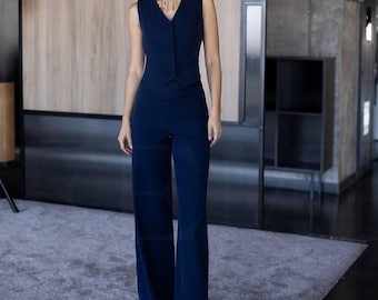 May include: A navy blue sleeveless vest with matching wide-leg pants. The vest has a V-neck and button closure. The pants are high-waisted and flared. The outfit is worn with a gold necklace. The background is a modern interior with wood paneling and a gray carpet.