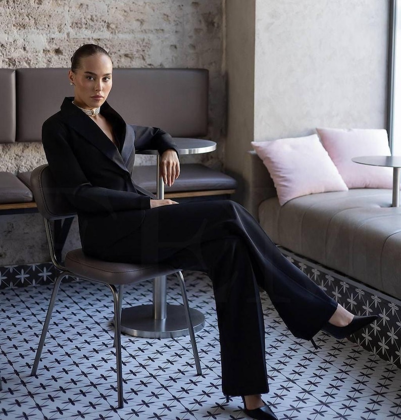 May include: A woman in a black suit sits on a chair. The suit includes a blazer and wide-leg pants. She wears black heels and a pearl necklace. The background features a cafe setting with a patterned tile floor and a neutral color palette.