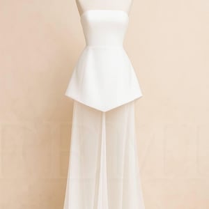 May include: A strapless white wedding dress on a mannequin. The bodice is fitted, and the skirt features a unique geometric overlay that flows into a sheer, floor-length train. The dress is made of a smooth, opaque fabric.