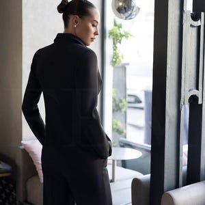 May include: A black suit, consisting of a blazer and matching pants. The blazer has a tailored fit with long sleeves and a notched lapel. The pants appear to be wide-legged. The suit is worn by a person standing near a window.