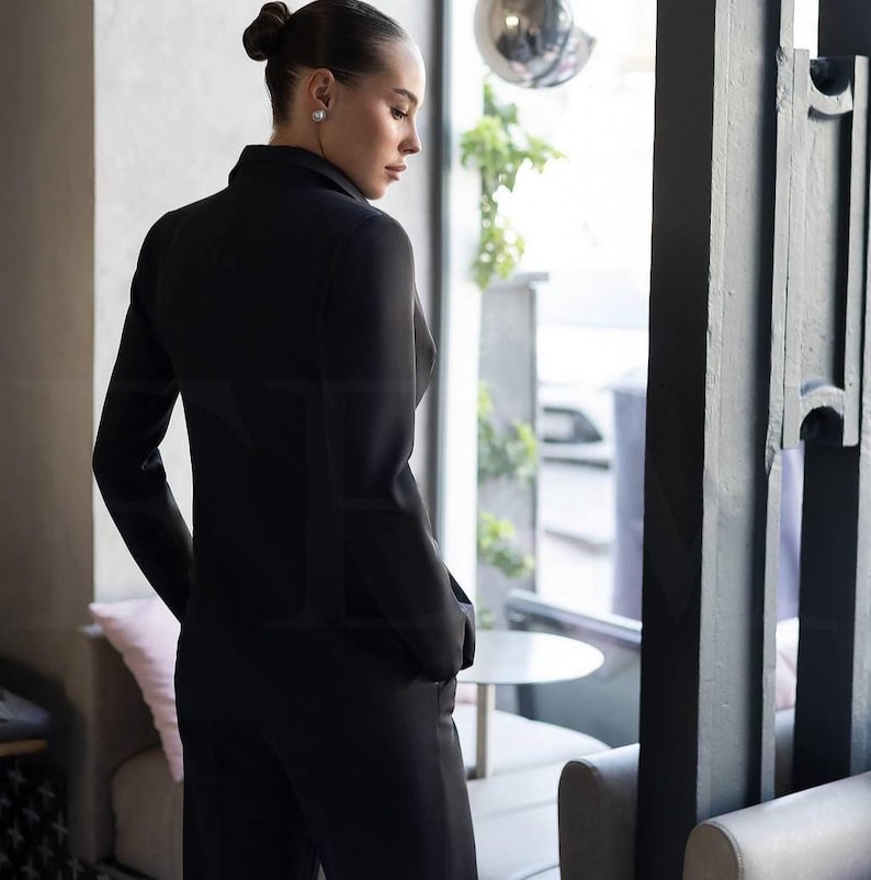 May include: A black suit, consisting of a blazer and matching pants. The blazer has a tailored fit with long sleeves and a notched lapel. The pants appear to be wide-legged. The suit is worn by a person standing near a window.