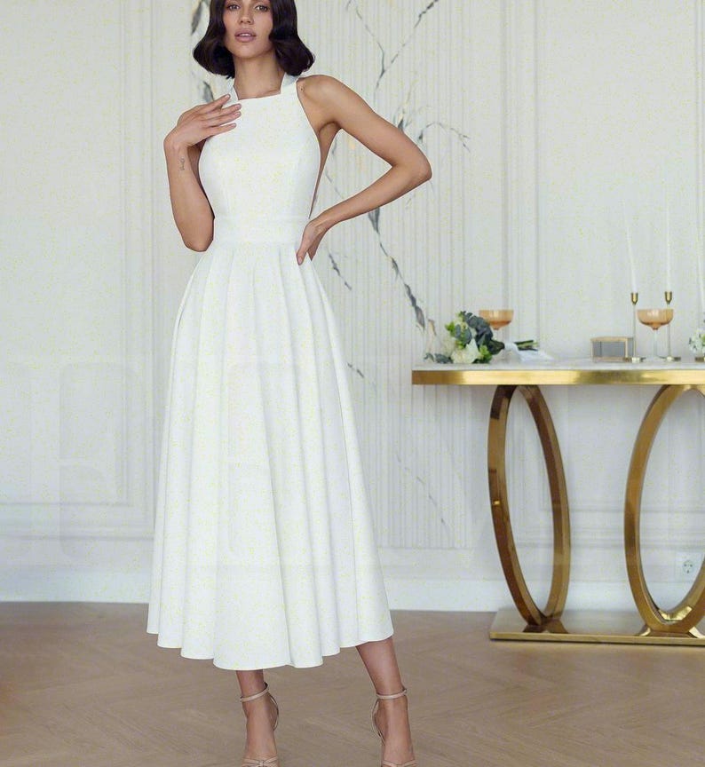 May include: A white halter-neck dress with a full skirt. The dress has a fitted bodice and a flared skirt that falls to mid-calf. The dress is paired with nude-colored heels. The background features a gold table and a marble-patterned wall.