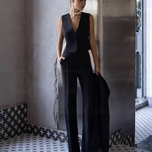 May include: A black sleeveless vest with a single button closure, paired with matching wide-leg trousers. A black blazer is held in the hand. The outfit is worn in a room with patterned tile flooring and a neutral-toned wall.