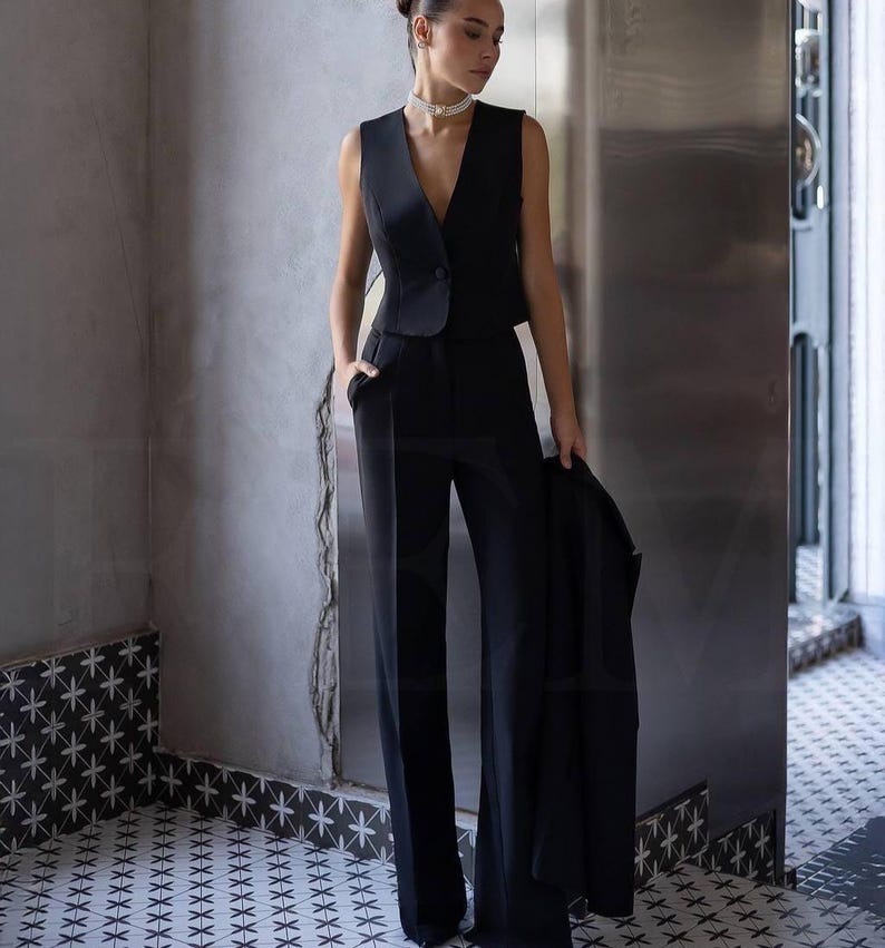 May include: A black sleeveless vest with a single button closure, paired with matching wide-leg trousers. A black blazer is held in the hand. The outfit is worn in a room with patterned tile flooring and a neutral-toned wall.