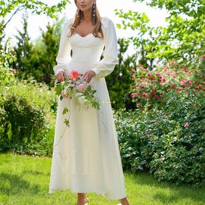 Pu&ograve; includere: Un abito da sposa bianco con maniche lunghe e scollatura a cuore. L'abito ha un corpetto aderente e una gonna fluida. La modella tiene un bouquet di fiori rosa e bianchi.