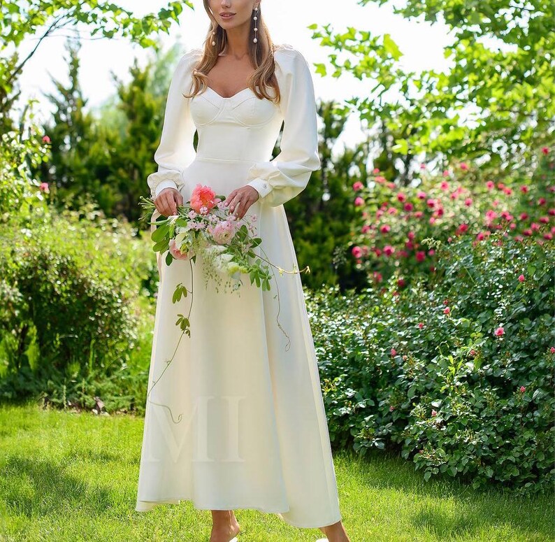 Pu&ograve; includere: Un abito da sposa bianco con maniche lunghe e scollatura a cuore. L'abito ha un corpetto aderente e una gonna fluida. La modella tiene un bouquet di fiori rosa e bianchi.