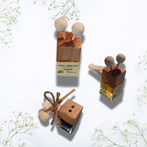 Op de afbeelding: Drie houten autoluchtverfrissers met verschillende ontwerpen. De eerste is een vierkante houten blok met twee houten ballen bovenop en een label met de tekst "Lemon & Bergamot Car Diffuser". De tweede is een houten blok met twee houten ballen bovenop en een heldere glazen fles met een gouden label. De derde is een houten blok met een heldere glazen fles en een enkele houten bal bovenop.