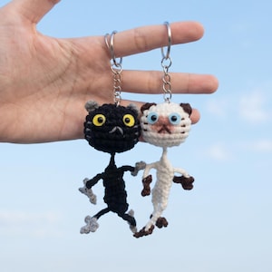 May include: Two handmade crochet cat keychains. One is black with yellow eyes, the other is white with brown accents. Both have silver key rings and are held against a blue sky background.