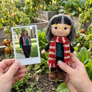 Puede incluir: Muñeca de ganchillo hecha a mano con pelo gris, gafas amarillas y bufanda roja y blanca. La muñeca sostiene una foto de una mujer con una bufanda similar. La muñeca viste un top negro, un suéter verde y pantalones negros.