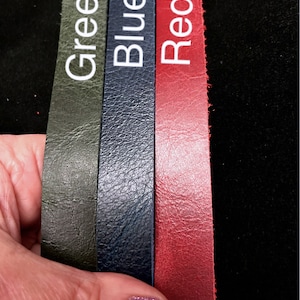 May include: Three leather strips in green, blue, and red are displayed. Each strip has a white label with its respective color name. The leather has a textured surface. The background is black.