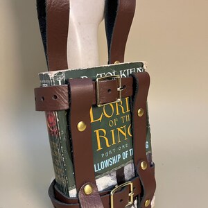 May include: A brown leather book holder with brass buckles and rivets, designed to carry a copy of "The Lord of the Rings: The Fellowship of the Ring." The book is visible within the holder.