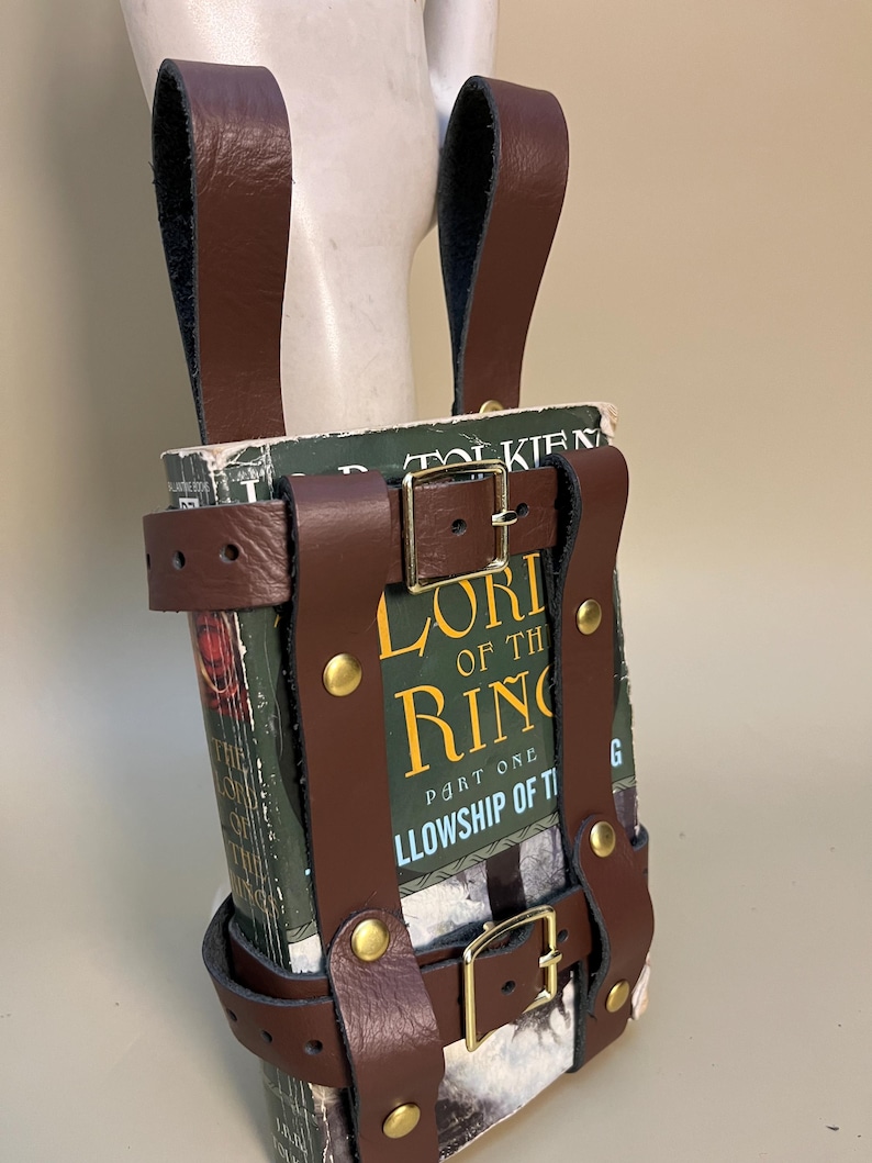 May include: A brown leather book holder with brass buckles and rivets, designed to carry a copy of "The Lord of the Rings: The Fellowship of the Ring." The book is visible within the holder.