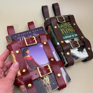 May include: Two leather book holders in burgundy and brown, secured with buckles and rivets. One holds a book with a woman in a red dress on the cover, the other holds a book with the title "Lord of the Rings".