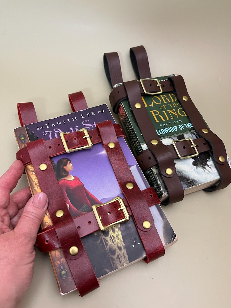 May include: Two leather book holders in burgundy and brown, secured with buckles and rivets. One holds a book with a woman in a red dress on the cover, the other holds a book with the title "Lord of the Rings".