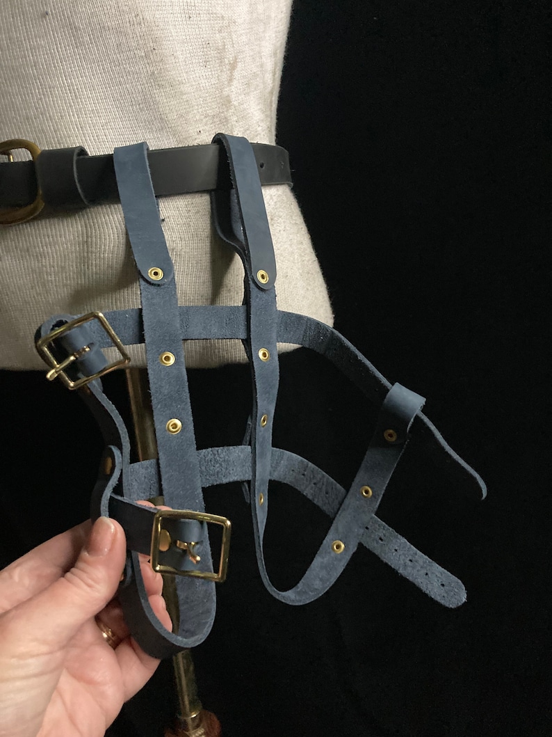 May include: A blue suede harness with brass buckles and rivets. The harness is attached to a black belt with a brass buckle. The harness is designed to be worn around the waist and legs.