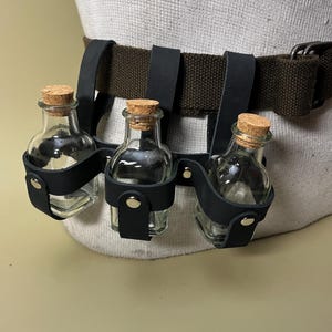 May include: A black leather belt with three small, clear glass bottles. Each bottle has a cork stopper. The bottles are held in place by black leather straps attached to the belt. The belt is brown canvas.