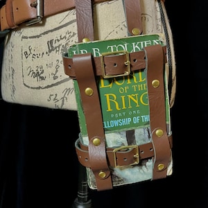May include: A brown leather belt with a book holder. The holder is secured with straps and brass buckles. The book cover reads "The Lord of the Rings: Part One, Fellowship of the Ring."