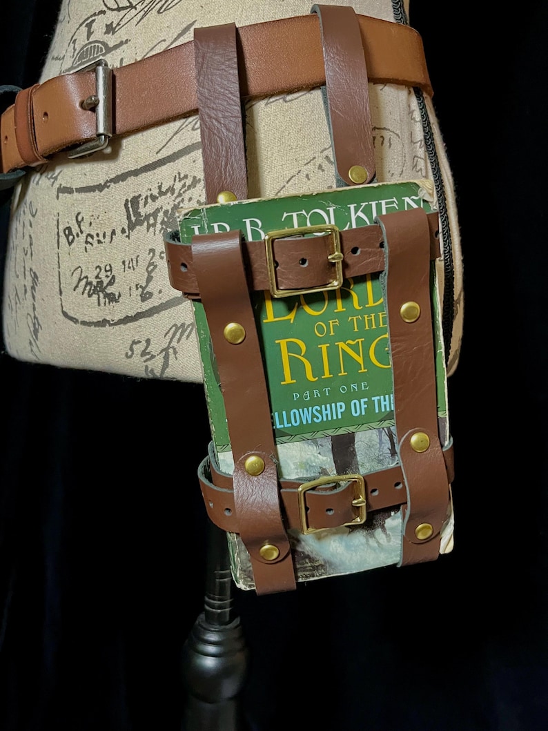 May include: A brown leather belt with a book holder. The holder is secured with straps and brass buckles. The book cover reads "The Lord of the Rings: Part One, Fellowship of the Ring."