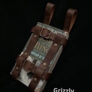 May include: A brown leather book carrier with buckles and straps, holding a copy of "The Lord of the Rings: The Fellowship of the Ring." The carrier has two loops for carrying. The word "Grizzly" is in the bottom right corner.